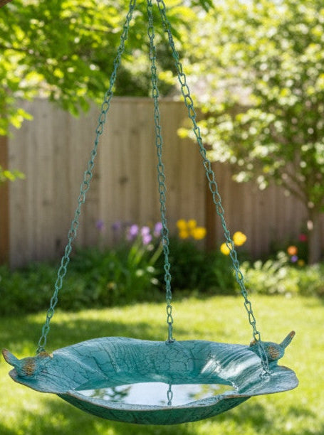 Teal bird bath hanging from a tree in a garden with flowers and grass.